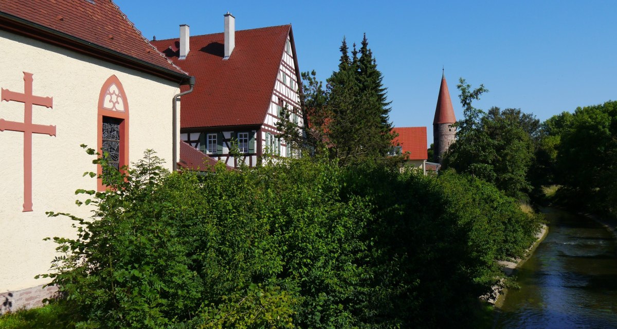 Fachwerkhaus und Kirche mit Turm an einem Flussufer, umgeben von Bäumen und blauem Himmel., © Natur.Nah. Schönbuch & Heckengäu