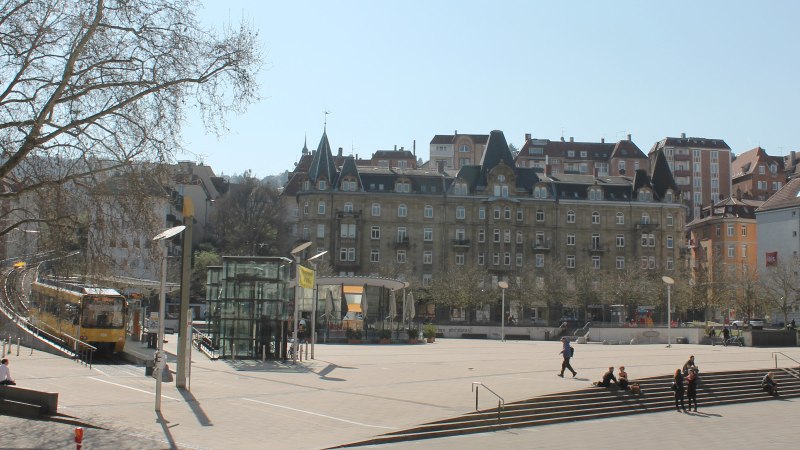 Marienplatz in Stuttgart mit Straßenbahn, modernen und historischen Gebäuden. Menschen sitzen auf Treppen, Bäume ohne Laub, sonniger Tag., © Stuttgart-Marketing GmbH