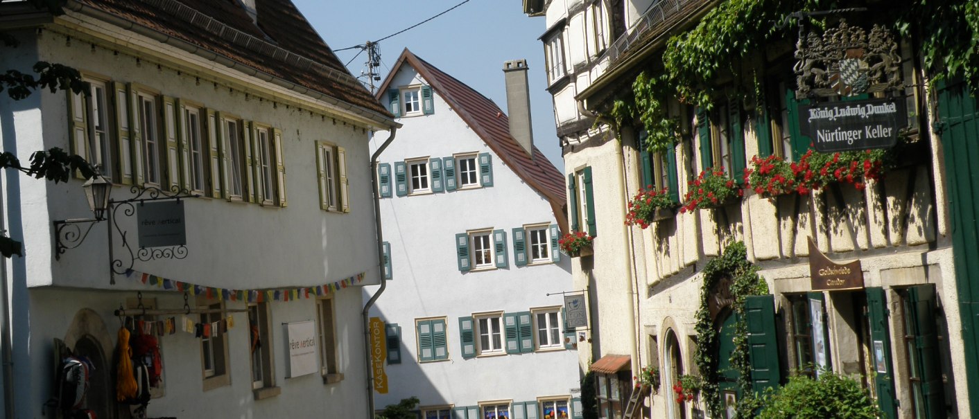 Malerische Altstadtgasse mit Fachwerkhäusern, blühenden Blumenkästen und einem Schild mit der Aufschrift 'Nürtinger Keller'., © Nürtingen Malerische Altstadtgasse mit Fachwerkhäusern, blühenden Blumenkästen und einem Schild mit der Aufschrift 'Nürtinger Keller'., © Nürtingen