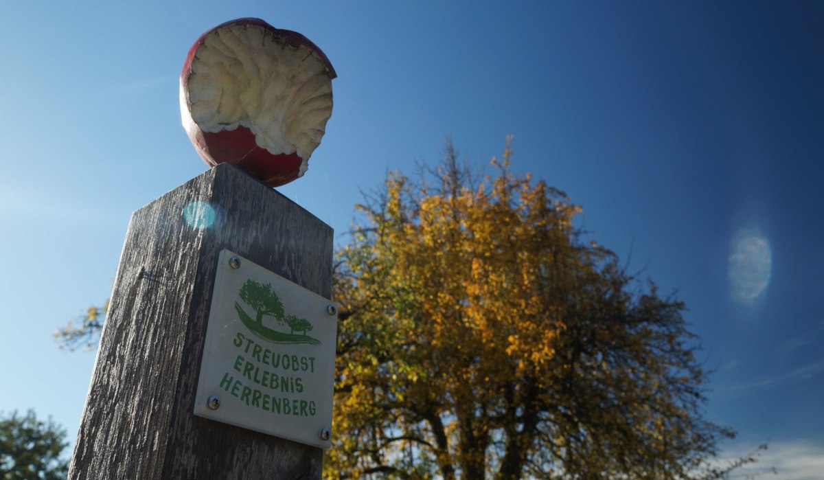 Ein Holzpfosten mit einem angebissenen Apfel und einem Schild 'Streuobst Erlebnis Herrenberg' vor einem Baum und blauem Himmel., &copy; Natur.Nah. Sch&ouml;nbuch & Heckeng&auml;u