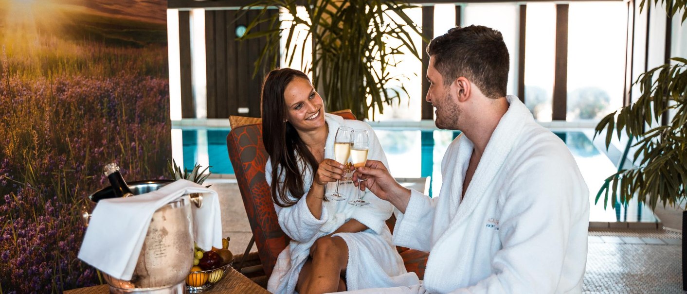 Ein Paar in weißen Bademänteln stößt mit Sektgläsern an. Sie sitzen auf Liegen, im Hintergrund ist ein Pool und Pflanzen zu sehen., © Ringhotel Adler Asperg