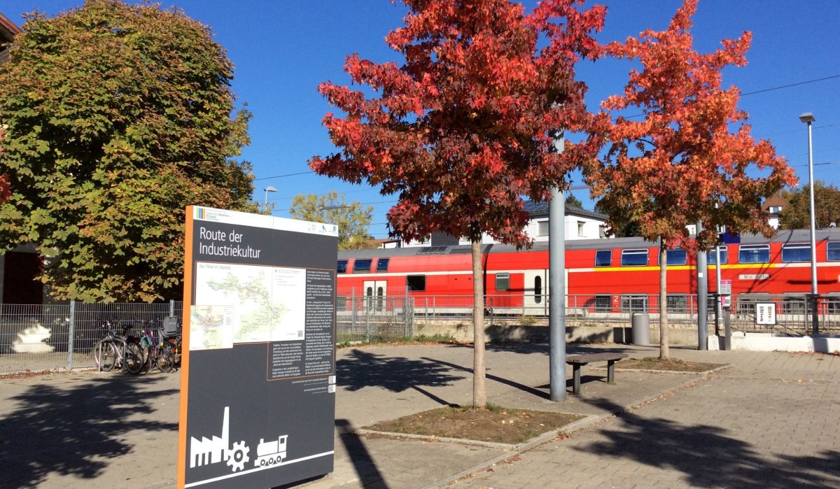 Schild der Route der Industriekultur neben roten Bäumen und einem vorbeifahrenden Zug. Herbstliche Atmosphäre mit klarem Himmel., © Herde