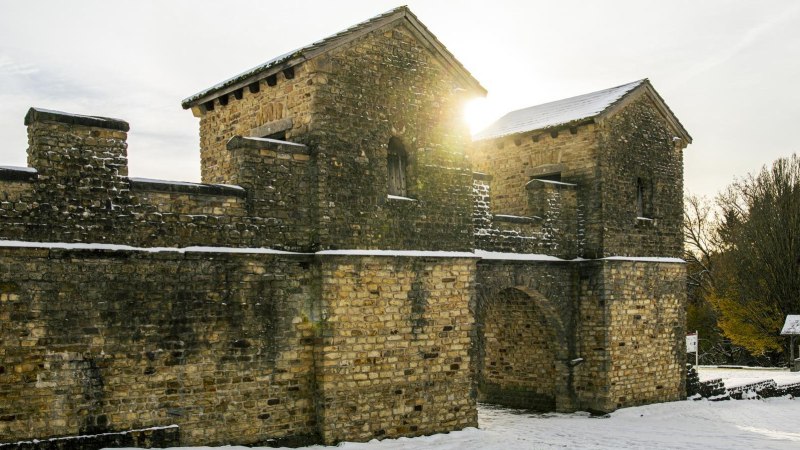 Ein römisches Kastell aus Stein im Schnee, beleuchtet von Sonnenstrahlen. Im Hintergrund sind Bäume und ein kleiner Pavillon zu sehen., © SMG, Sarah Schmid Ein römisches Kastell aus Stein im Schnee, beleuchtet von Sonnenstrahlen. Im Hintergrund sind Bäume und ein kleiner Pavillon zu sehen., © SMG, Sarah Schmid