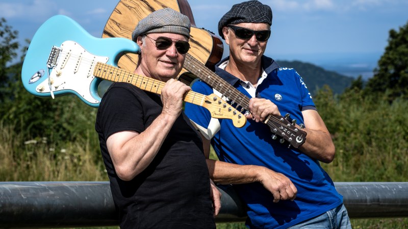 Zwei M&auml;nner mit Gitarren posieren l&auml;ssig im Freien. Sie tragen Sonnenbrillen und M&uuml;tzen, im Hintergrund ist eine h&uuml;gelige Landschaft zu sehen., &copy; (c) G&uuml;nter Orban