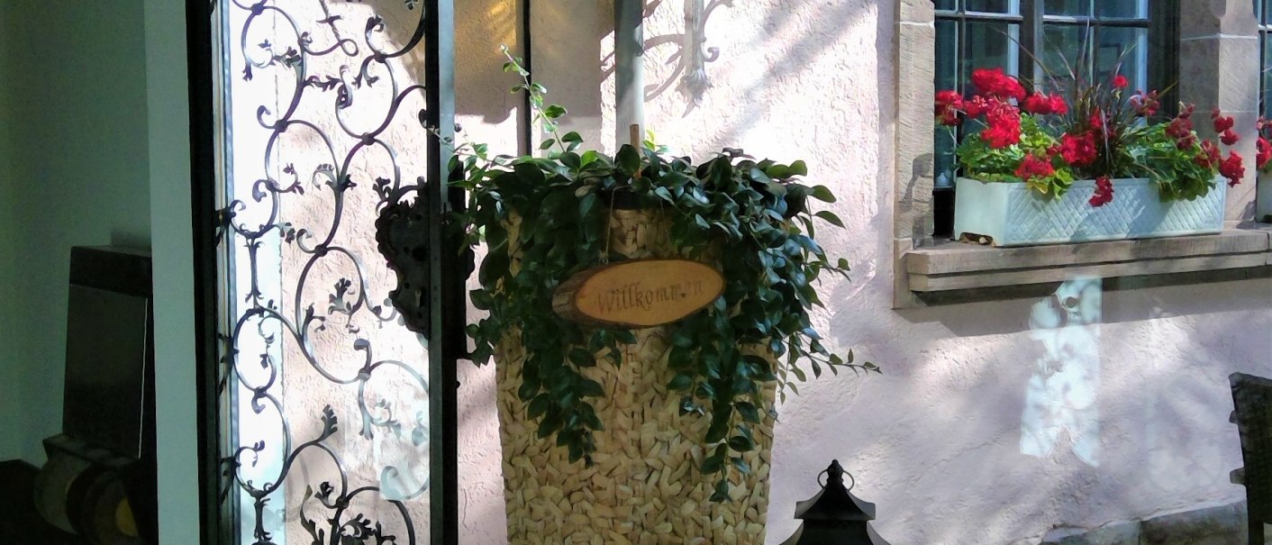 Ein einladender Eingang mit einem offenen schmiedeeisernen Tor, einem großen Pflanzenkorb mit 'Willkommen'-Schild, Laternen und einem Blumenkasten am Fenster., © relexa Waldhotel Schatten Ein einladender Eingang mit einem offenen schmiedeeisernen Tor, einem großen Pflanzenkorb mit 'Willkommen'-Schild, Laternen und einem Blumenkasten am Fenster., © relexa Waldhotel Schatten