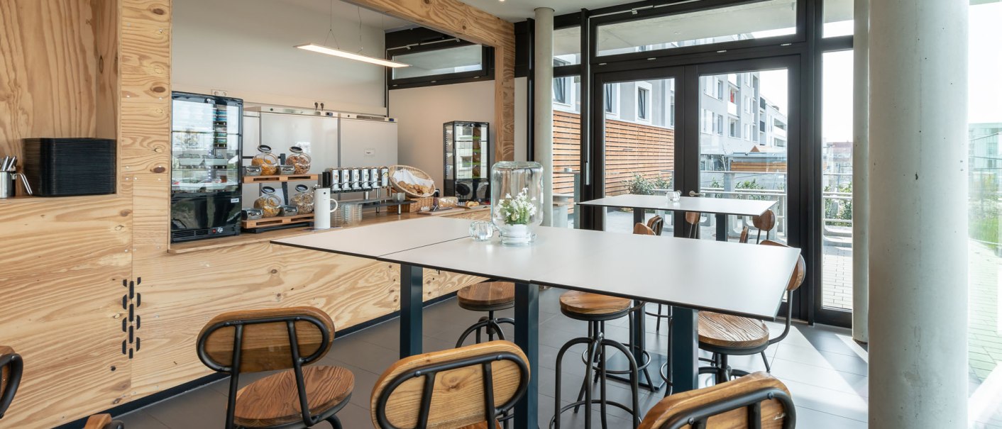 Moderner Frühstücksbereich mit hohen Tischen und Stühlen aus Holz. Große Fenster lassen viel Licht herein. Im Hintergrund ein Buffet mit Getränken und Gebäck., © a2 HOTELS