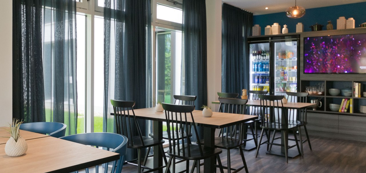 Moderner Raum mit Holztischen, schwarzen und blauen Stühlen. Kühlschrank mit Getränken, Fernseher an der Wand. Dunkle Vorhänge und Holzboden., © Arthotel Ana / elaya