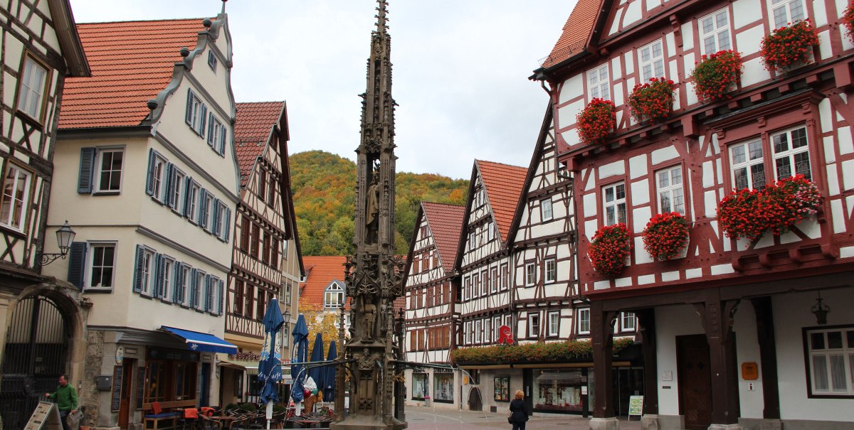 Ein malerischer Marktplatz mit Fachwerkhäusern, einem verzierten Brunnen und herbstlich gefärbten Bäumen im Hintergrund.