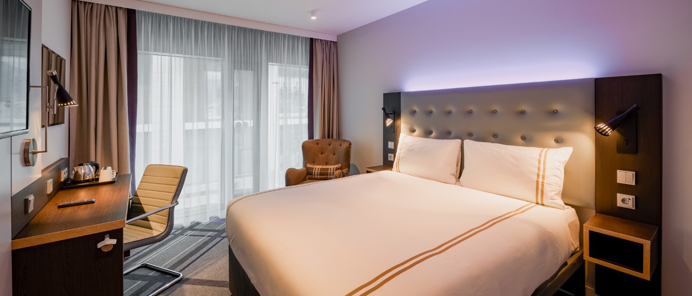 Modernes Doppelzimmer mit großem Bett, Schreibtisch, Sessel und Fenster mit Vorhängen. Dezente Beleuchtung und stilvolle Einrichtung., © Premier Inn