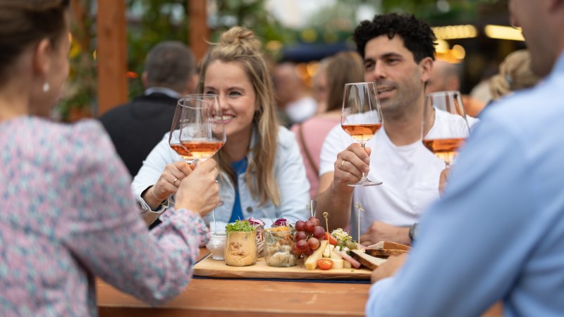 Vier Personen sto&szlig;en mit Weingl&auml;sern an einem Tisch im Freien an, umgeben von Essen und einer geselligen Atmosph&auml;re., &copy; Stadt N&uuml;rtingen, B. Knoblauch