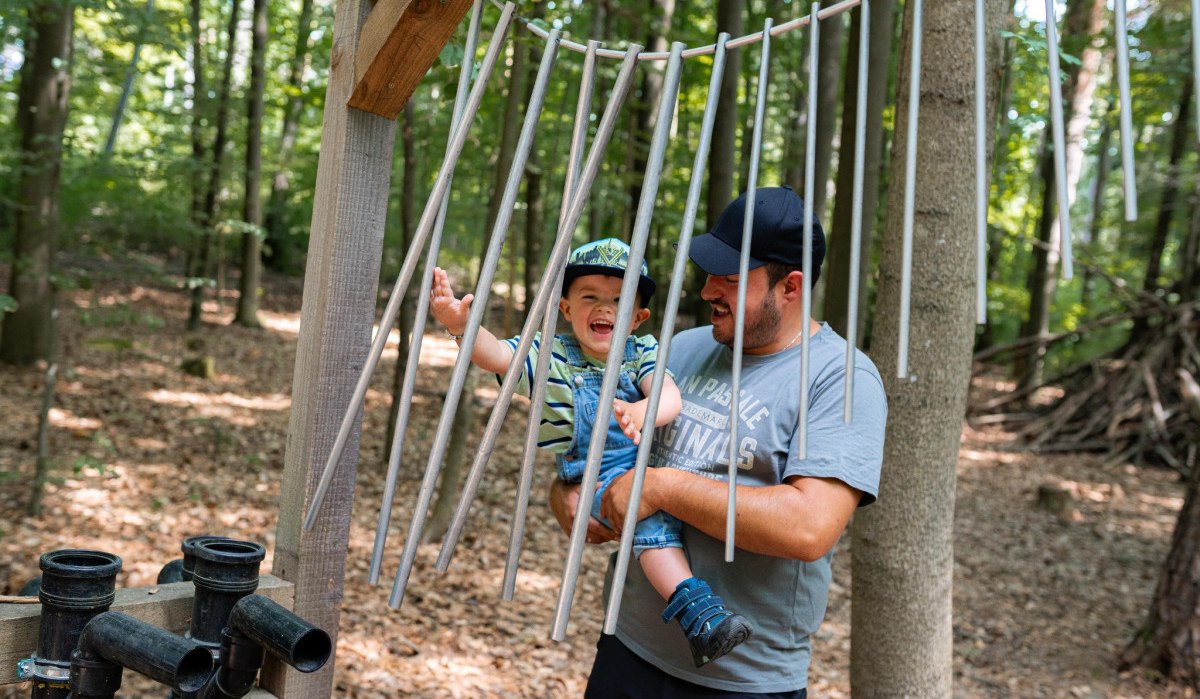 Ein Mann hält ein lachendes Kind in einem Wald. Sie stehen zwischen Metallrohren, die an einem Holzrahmen befestigt sind., © Stadtmarketing Weil der Stadt Ein Mann hält ein lachendes Kind in einem Wald. Sie stehen zwischen Metallrohren, die an einem Holzrahmen befestigt sind., © Stadtmarketing Weil der Stadt