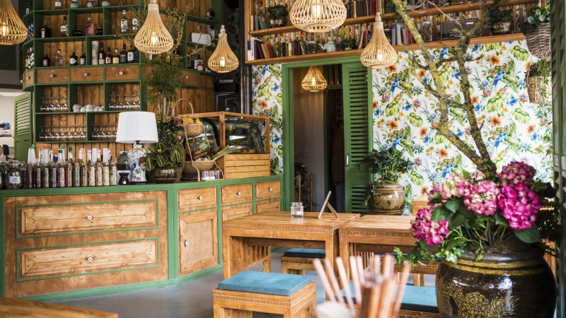 Gemütlicher Gastraum mit Holzmöbeln, bunten Blumen, Regalen voller Flaschen und Büchern. Helle Lampen und florale Tapete schaffen eine einladende Atmosphäre., © Vietal Kitchen