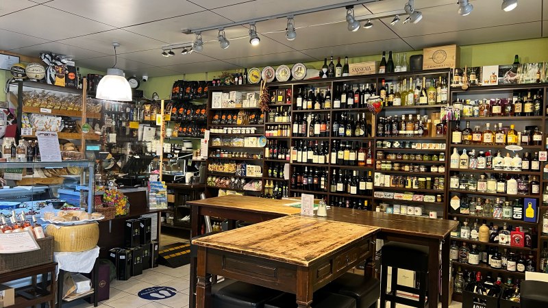 Feinkostladen mit Regalen voller Wein, Spirituosen und Delikatessen. Ein Holztisch mit St&uuml;hlen steht im Vordergrund. Der Raum ist hell beleuchtet., &copy; Feinkost-Galeria