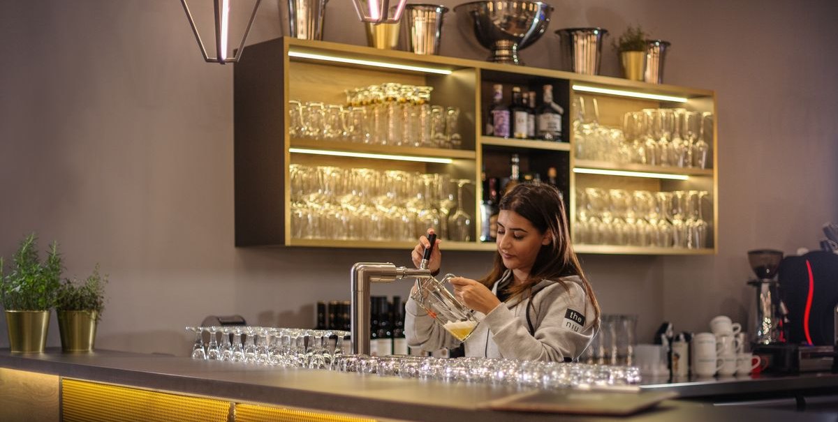 Frau zapft Bier an einer modernen Bar mit beleuchteten Regalen und Gläsern. Elegante Beleuchtung und Dekoration., © Novum Hospitality Frau zapft Bier an einer modernen Bar mit beleuchteten Regalen und Gläsern. Elegante Beleuchtung und Dekoration., © Novum Hospitality