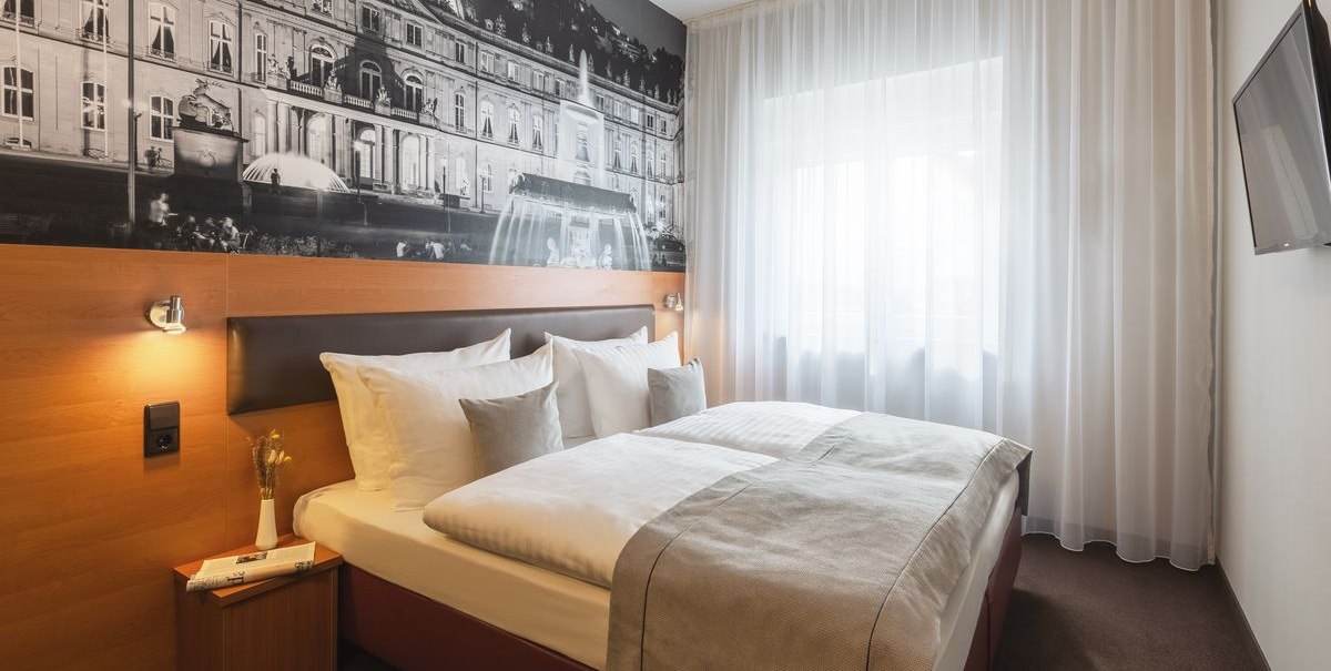 Modernes Hotelzimmer mit Doppelbett, großem Wandbild eines Gebäudes, Fenster mit weißem Vorhang und Wand-TV., © Novum Hospitality