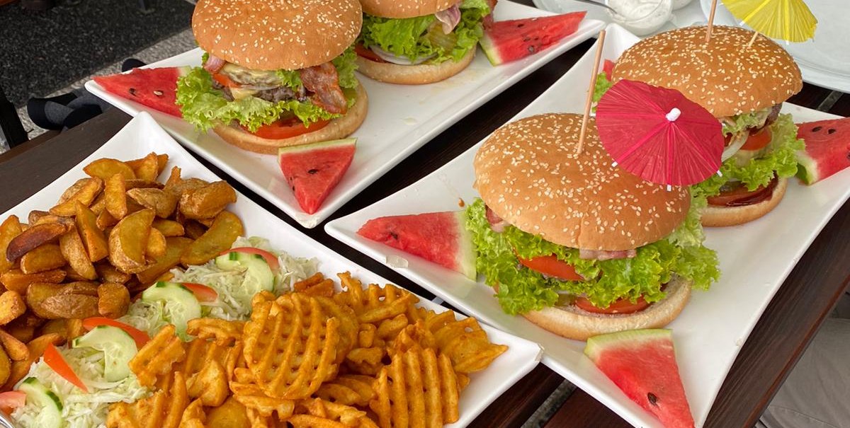 Drei große Burger mit bunten Schirmchen, serviert mit Pommes und Salat auf einem Tisch. Im Hintergrund ein Getränk und eine Person in kariertem Hemd., © Gaststätte Goldbachsee Drei große Burger mit bunten Schirmchen, serviert mit Pommes und Salat auf einem Tisch. Im Hintergrund ein Getränk und eine Person in kariertem Hemd., © Gaststätte Goldbachsee