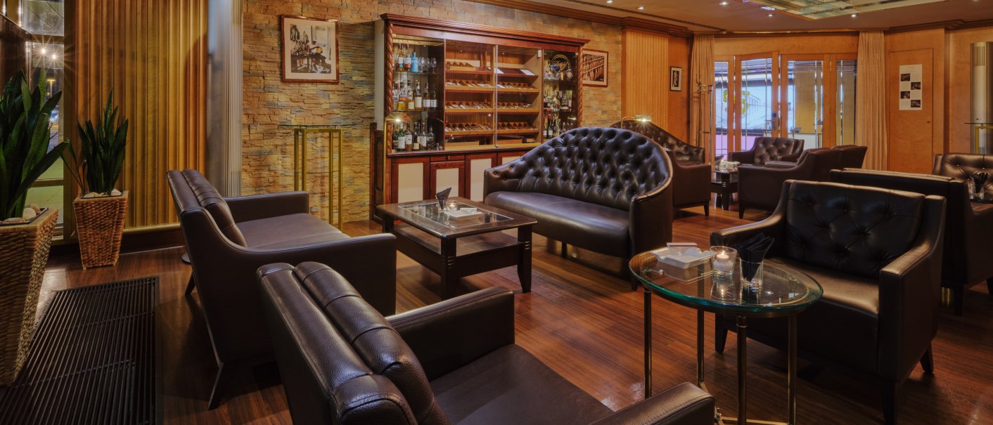 Elegante Lounge mit Ledersesseln, Holzboden, Pflanzen und einem Humidor im Hintergrund. Dezente Beleuchtung und stilvolle Einrichtung., © Steigenberger Graf Zeppelin