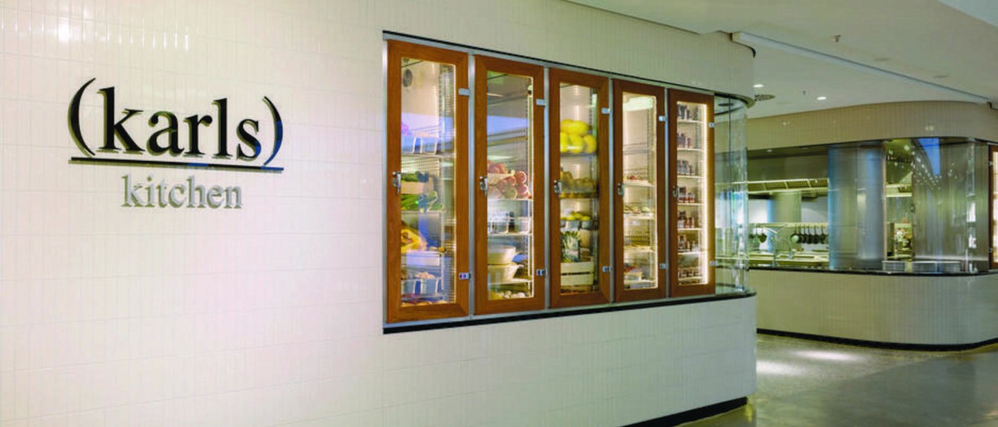 Eingang zu "karls kitchen" mit beleuchteten Glasvitrinen voller Lebensmittel und Blick in die moderne Küche., © karls kitchen, Stuttgart Eingang zu "karls kitchen" mit beleuchteten Glasvitrinen voller Lebensmittel und Blick in die moderne Küche., © karls kitchen, Stuttgart