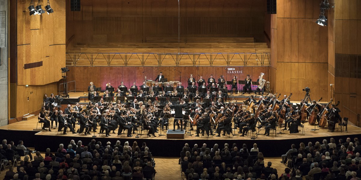 Das SWR Symphonieorchester spielt auf einer gro&szlig;en B&uuml;hne vor Publikum. Die Musiker tragen formelle Kleidung, und der Dirigent steht in der Mitte., &copy; SWR Alexander Kluge