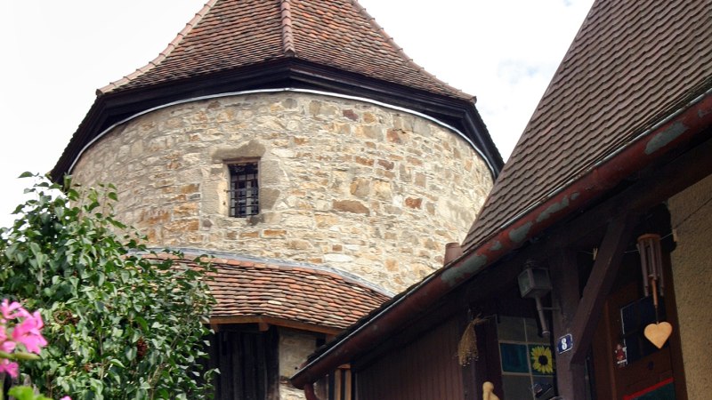 Ein mittelalterlicher Steinturm mit spitzem Dach erhebt sich über Fachwerkhäusern, umgeben von blühenden Blumen und blauem Himmel., © Mende Ein mittelalterlicher Steinturm mit spitzem Dach erhebt sich über Fachwerkhäusern, umgeben von blühenden Blumen und blauem Himmel., © Mende
