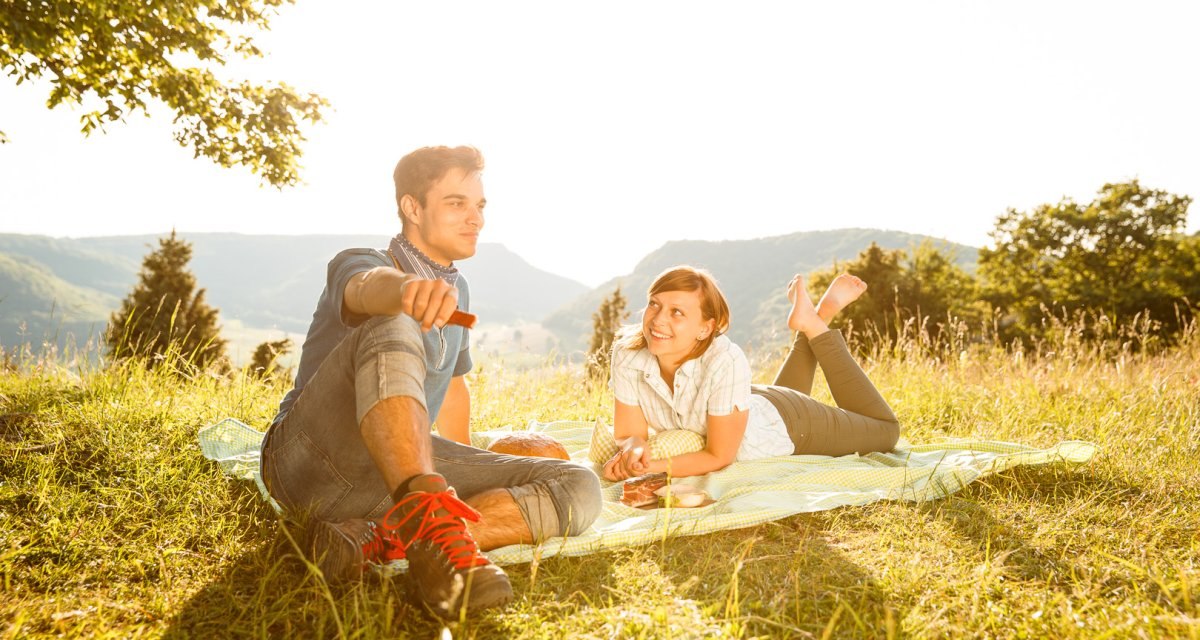 Ein Paar sitzt auf einer Decke bei einem Picknick auf einer sonnigen Wiese mit Bergblick., © Distretto di Göppingen Ein Paar sitzt auf einer Decke bei einem Picknick auf einer sonnigen Wiese mit Bergblick., © Distretto di Göppingen
