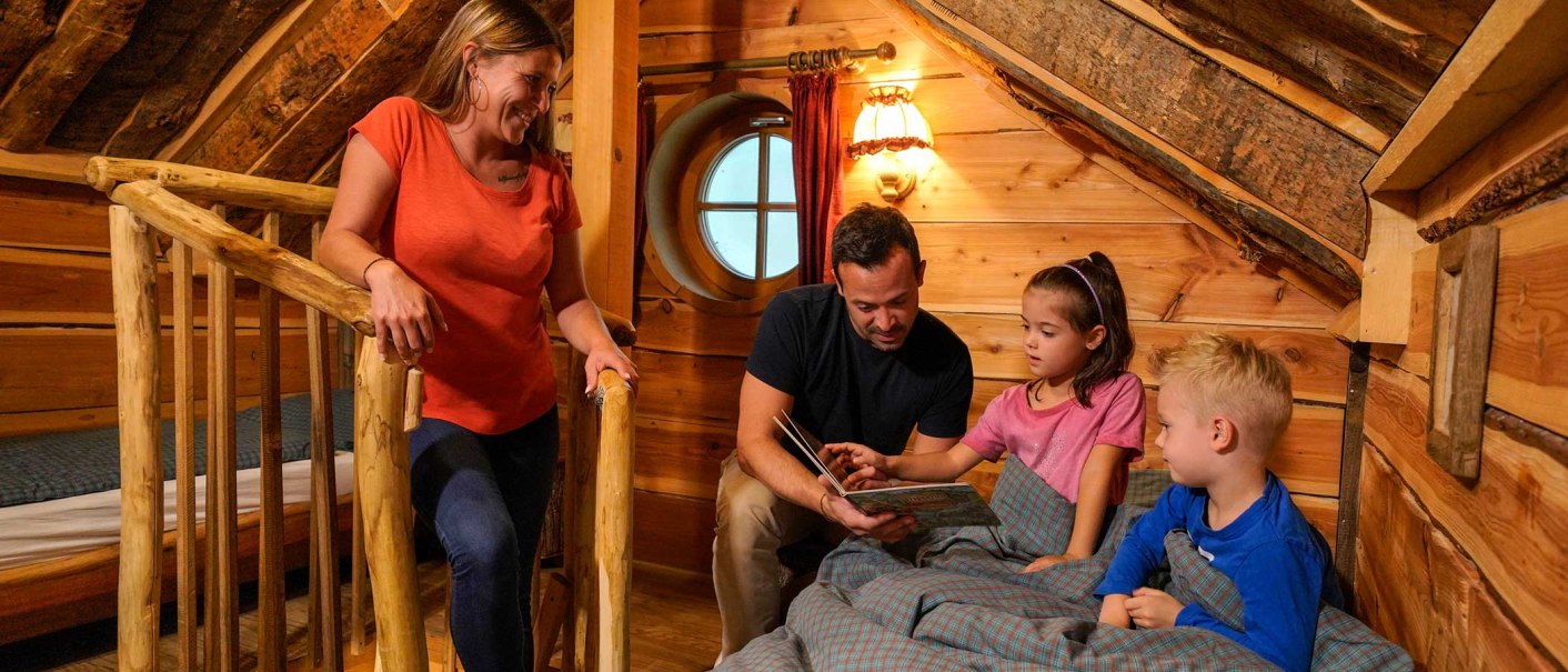 Eine Familie in einem gemütlichen Baumhaus. Der Vater liest den Kindern vor, während die Mutter lächelnd zusieht. Holzwände und rustikale Einrichtung., © Tripsdrill Natur-Resort GmbH