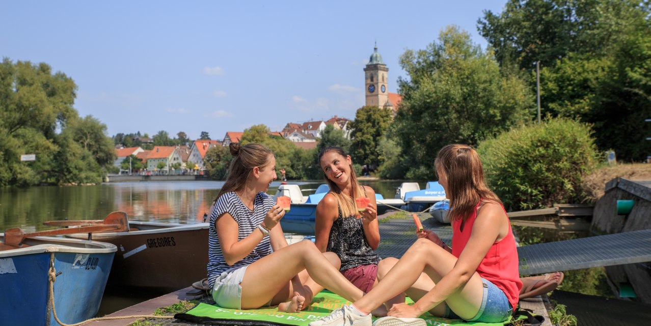 Drei Frauen sitzen auf einem Steg am Fluss und essen Wassermelone. Im Hintergrund sind Boote und ein Kirchturm zu sehen., © H. Bergmüller Drei Frauen sitzen auf einem Steg am Fluss und essen Wassermelone. Im Hintergrund sind Boote und ein Kirchturm zu sehen., © H. Bergmüller