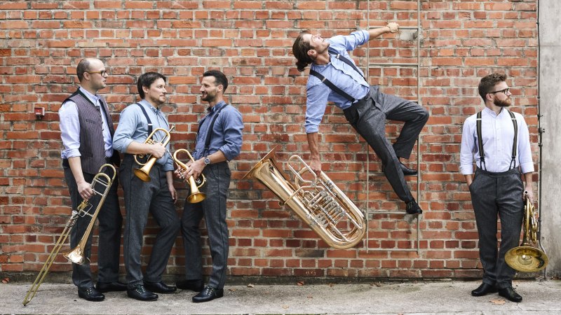 F&uuml;nf junge M&auml;nner in legerer Kleidung vor einer Wand aus Ziegelsteinen mit Blasinstrumenten in den H&auml;nden.  Zweiter von rechts klettert mit einer Tuba in der Hand  an einer Feuerleiter hinauf., &copy; munich brass connection