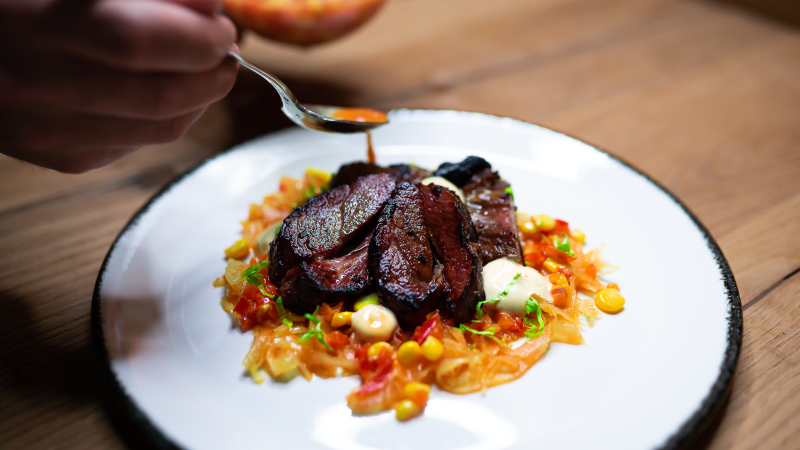 Ein kunstvoll angerichtetes Gericht mit Fleisch, Gemüse und Soße auf einem weißen Teller. Eine Hand hält einen Löffel über dem Teller., © Cara Schmid Ein kunstvoll angerichtetes Gericht mit Fleisch, Gemüse und Soße auf einem weißen Teller. Eine Hand hält einen Löffel über dem Teller., © Cara Schmid