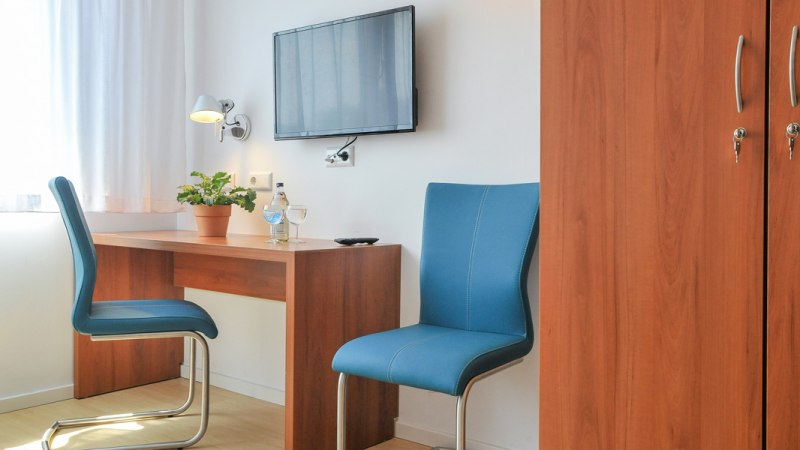 Ein schlichtes Hotelzimmer mit Schreibtisch, zwei blauen Stühlen, einem Fernseher an der Wand und einem Kleiderschrank., © Gästehaus ABZ Stuttgart Ein schlichtes Hotelzimmer mit Schreibtisch, zwei blauen Stühlen, einem Fernseher an der Wand und einem Kleiderschrank., © Gästehaus ABZ Stuttgart