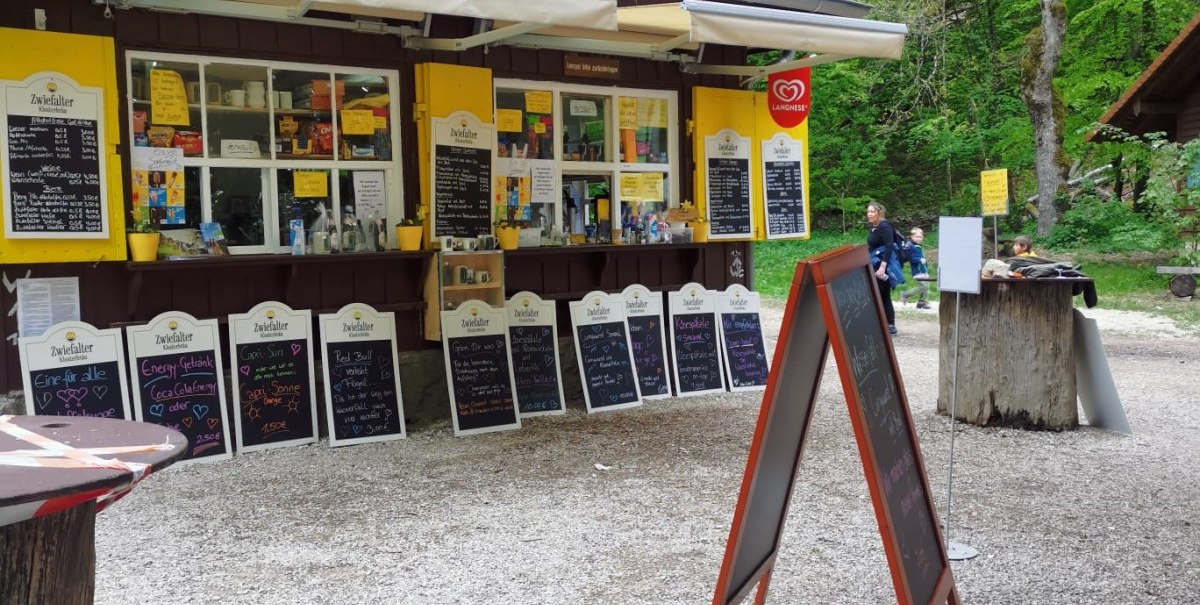 Kleiner Kiosk im Wald mit roten Dachziegeln, gelben Fensterläden und zahlreichen Schildern. Besucher im Hintergrund, umgeben von grüner Natur., © n/a