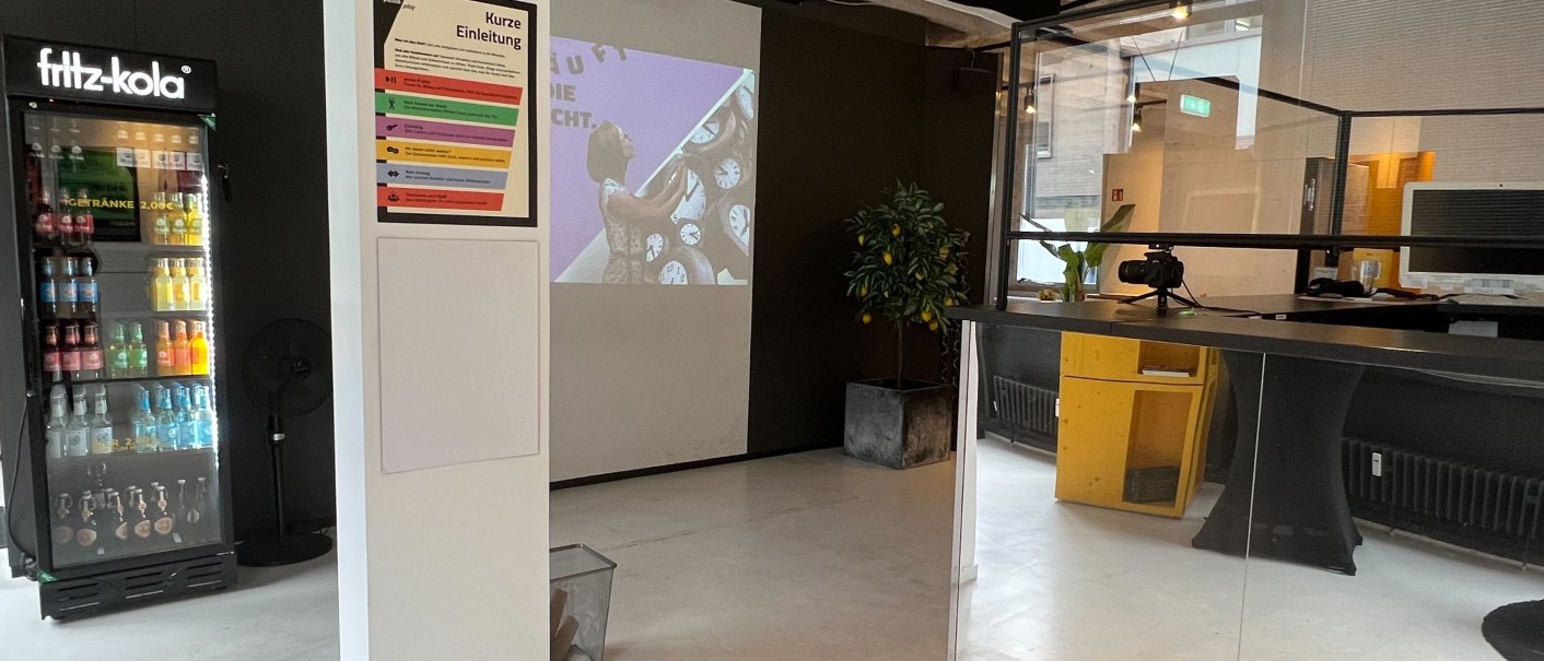 Ein Raum mit Kühlschrank, Projektor und Empfangstresen. An der Wand hängt ein Einleitungsposter. Der Raum wirkt modern und einladend., © pause & play Stuttgart