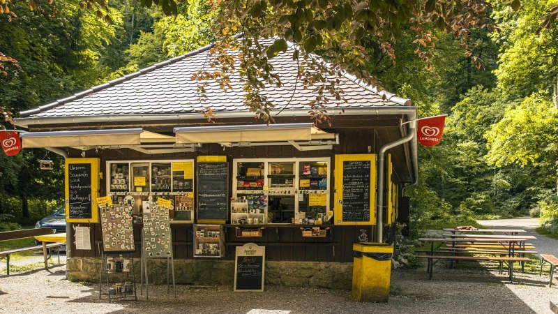 Ein Kiosk im Wald mit Verkaufstafeln und Sitzbänken. Langnese-Flaggen hängen an den Seiten. Der Weg führt durch die grüne Umgebung., © Stuttgart-Marketing GmbH, Sarah Schmid