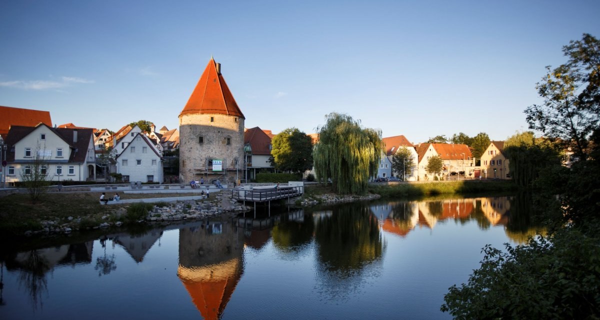 Ein mittelalterlicher Turm mit rotem Dach und umliegende Häuser spiegeln sich in einem ruhigen Fluss. Die Szene ist von Bäumen umgeben., © Stoppel Ein mittelalterlicher Turm mit rotem Dach und umliegende Häuser spiegeln sich in einem ruhigen Fluss. Die Szene ist von Bäumen umgeben., © Stoppel