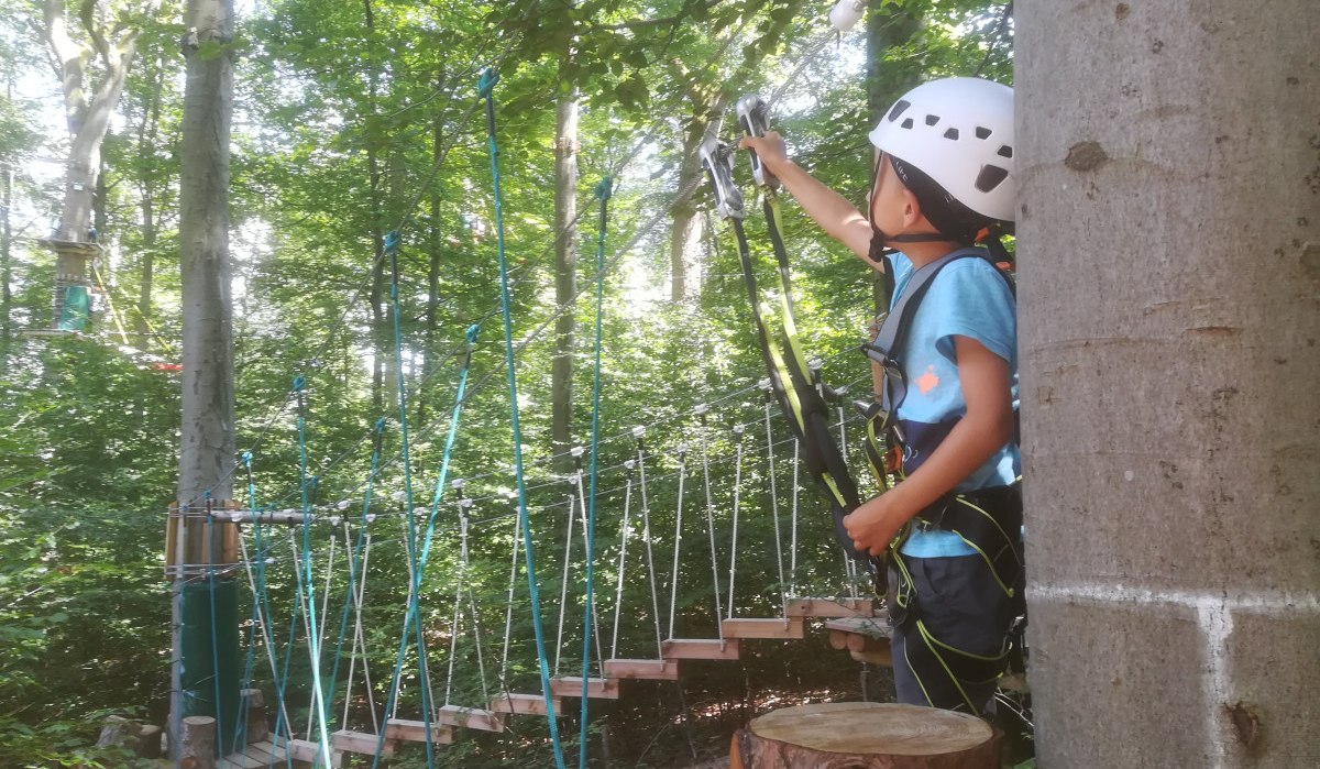 Ein Kind mit Helm und Kletterausrüstung steht in einem Kletterwald und greift nach einem Seil. Im Hintergrund sind Bäume und ein Kletterparcours zu sehen., © Kulturamt Plochingen Ein Kind mit Helm und Kletterausrüstung steht in einem Kletterwald und greift nach einem Seil. Im Hintergrund sind Bäume und ein Kletterparcours zu sehen., © Kulturamt Plochingen