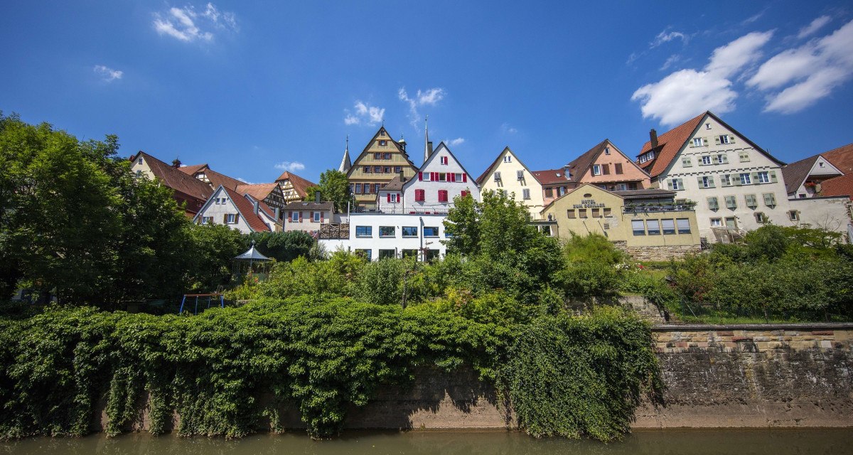 Bunte Fachwerkhäuser auf einem Hügel über einem Fluss, umgeben von Bäumen, unter blauem Himmel., © Stuttgart Marketing GmbH Achim Mende