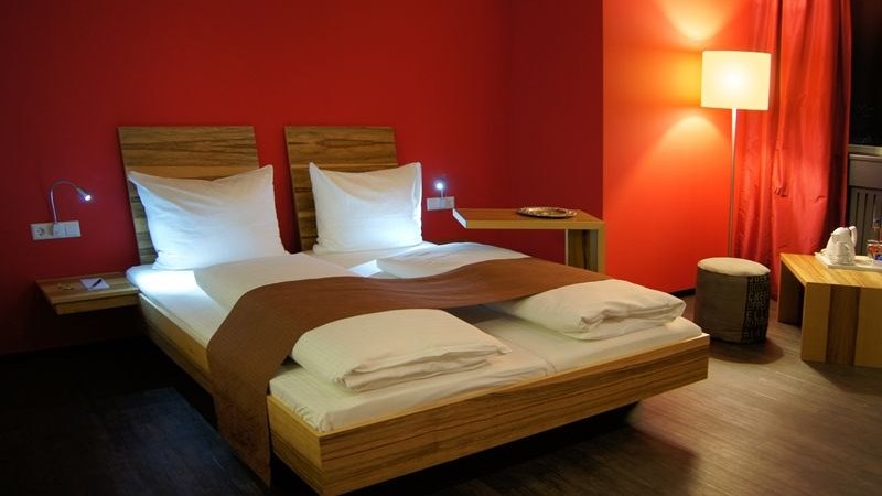 Modernes Hotelzimmer mit roter Wand, Doppelbett, Nachttischlampen und Stehlampe. Elegante Einrichtung mit Holzm&ouml;beln und warmem Licht., &copy; AMH Airport-Messe-Hotel