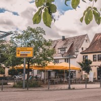 Gemütliche Straßenszene mit Fachwerkhäusern, gelben Sonnenschirmen und einem Café. Ein Wegweiser zeigt Richtungen an. Blätter hängen im Vordergrund., © Stadt Welzheim Gemütliche Straßenszene mit Fachwerkhäusern, gelben Sonnenschirmen und einem Café. Ein Wegweiser zeigt Richtungen an. Blätter hängen im Vordergrund., © Stadt Welzheim