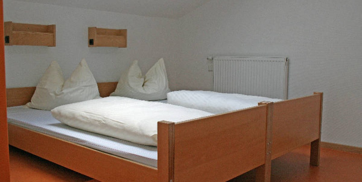 Ein schlichtes Zimmer mit zwei Einzelbetten, weißen Bettdecken und Kissen. Holzmöbel und ein Heizkörper an der Wand.