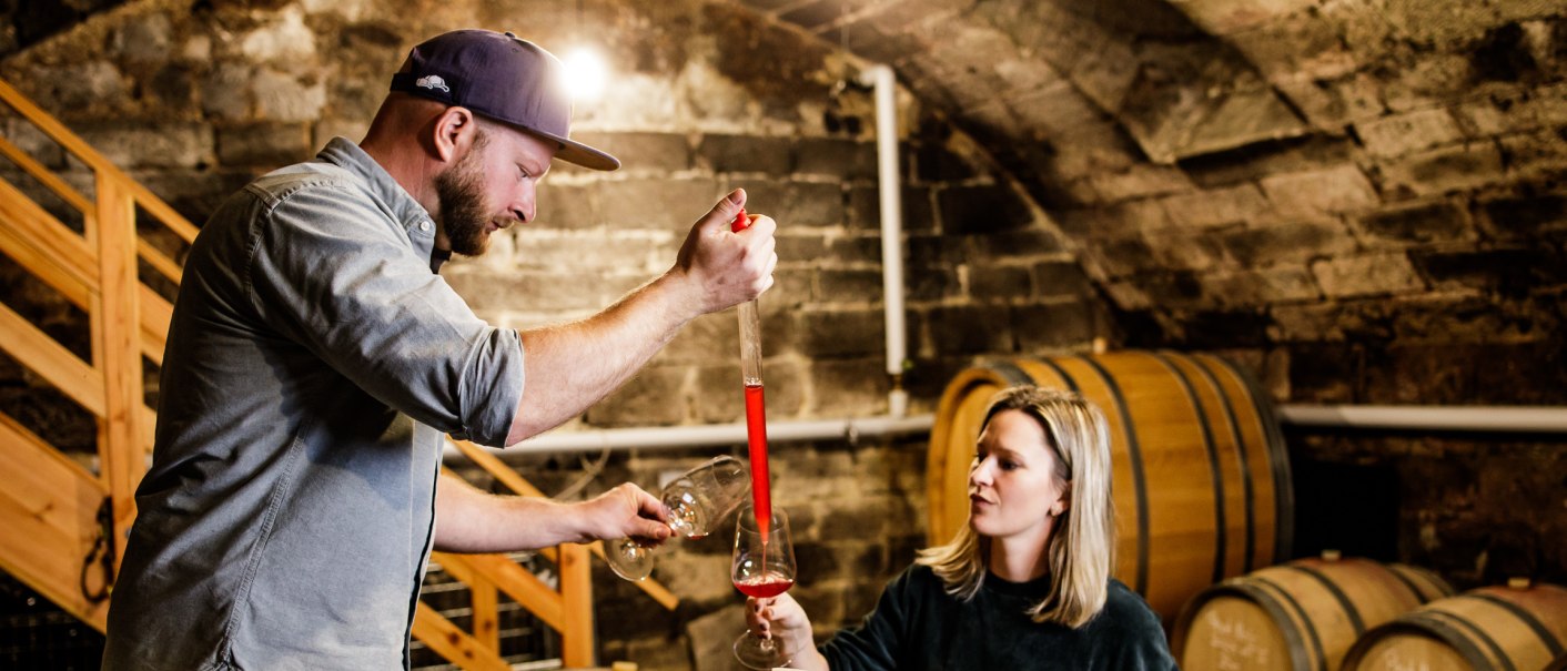 Zwei Personen im Weinkeller, die Wein aus einem Fass mit einem Glas probieren. Im Hintergrund sind Holzfässer zu sehen., © Weinbau Wieder Zwei Personen im Weinkeller, die Wein aus einem Fass mit einem Glas probieren. Im Hintergrund sind Holzfässer zu sehen., © Weinbau Wieder