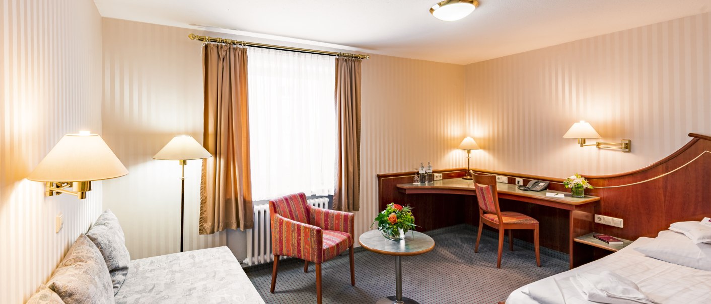 Gemütliches Hotelzimmer mit Bett, Sofa, Tisch und Stuhl. Beige gestreifte Wände, mehrere Lampen und ein Fenster mit Vorhängen., © TOMAS
