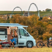 Wohnmobil mit Markise auf Wiese, zwei Personen am Tisch, Fahrr&auml;der daneben, Achterbahnen im Hintergrund, herbstliche Landschaft., &copy; Erlebnispark Tripsdrill GmbH & Co. KG