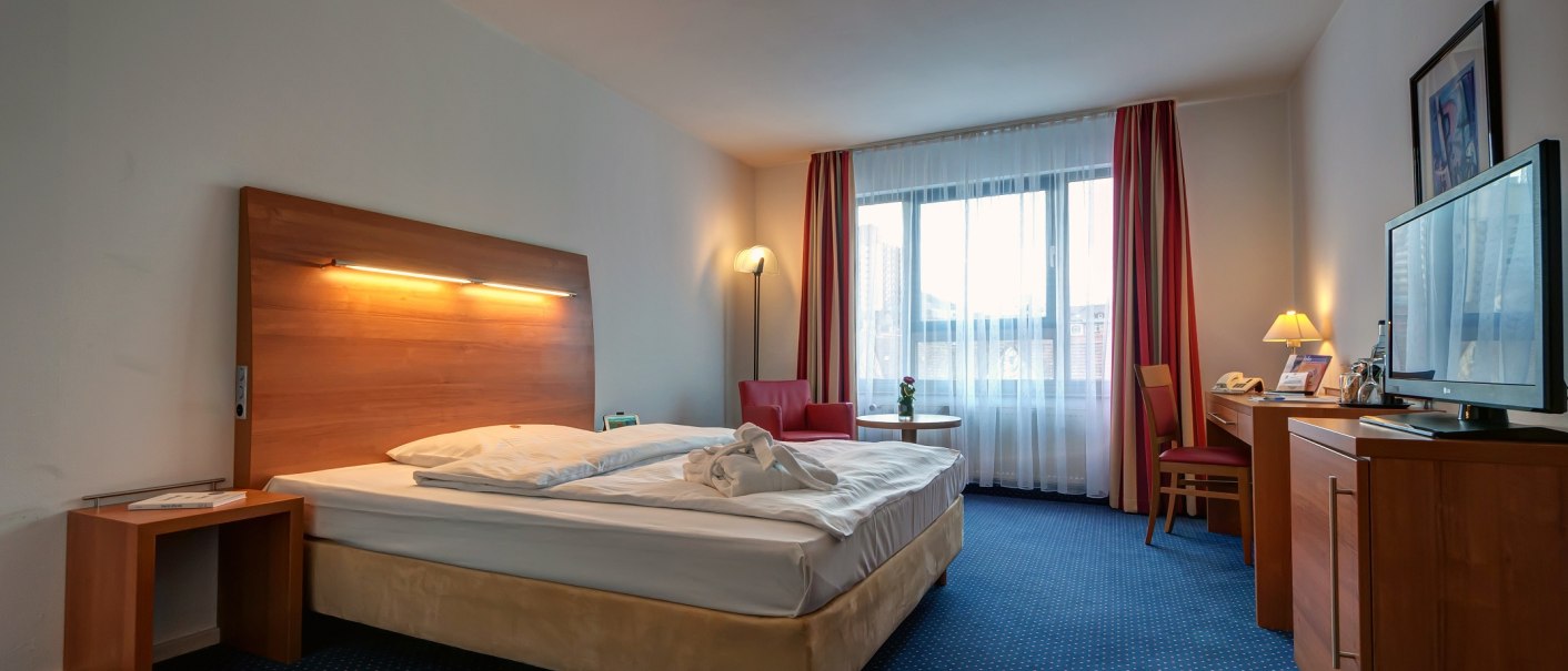 Ein gemütliches Hotelzimmer mit Doppelbett, Nachttisch, Schreibtisch, Fernseher und großem Fenster mit Vorhängen., © City Hotel Reutlingen