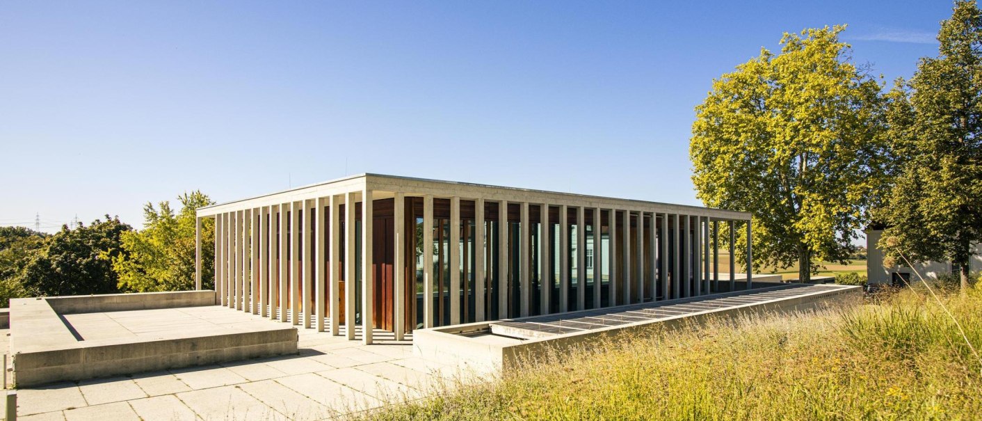 Modernes Gebäude mit Glasfassade und Säulen, umgeben von Bäumen und Wiese unter klarem Himmel., © SMG, Sarah Schmid Modernes Gebäude mit Glasfassade und Säulen, umgeben von Bäumen und Wiese unter klarem Himmel., © SMG, Sarah Schmid