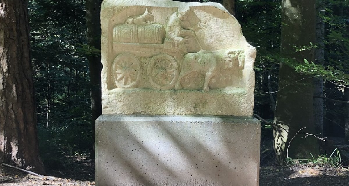 Ein Steinrelief zeigt einen Ochsenkarren und steht auf einem Sockel im Wald. Sonnenlicht fällt durch die Bäume auf das Relief., © Foto Julia Schaumann Ein Steinrelief zeigt einen Ochsenkarren und steht auf einem Sockel im Wald. Sonnenlicht fällt durch die Bäume auf das Relief., © Foto Julia Schaumann