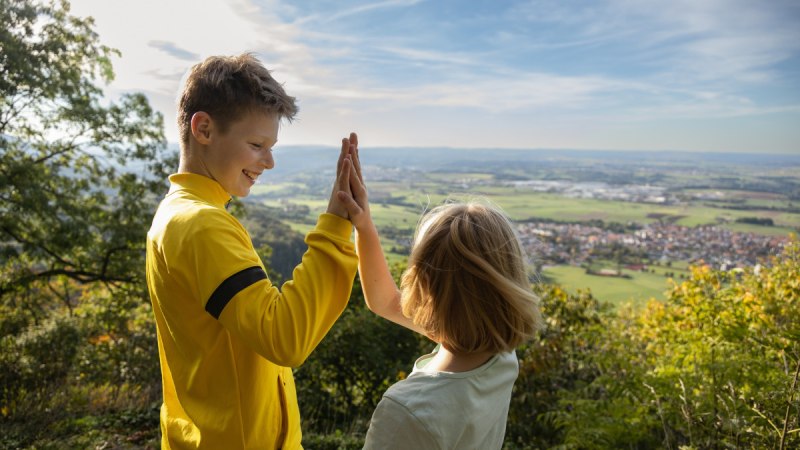 Zwei Kinder geben sich ein High-Five auf einem H&uuml;gel mit Blick auf eine weite Landschaft und einen blauen Himmel., &copy; Foto Thomas Zehnder