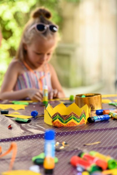 Ein Kind bastelt konzentriert an einem Tisch. Im Vordergrund liegt eine bunte Papierkrone, umgeben von Bastelmaterialien wie Klebestiften und Papier.