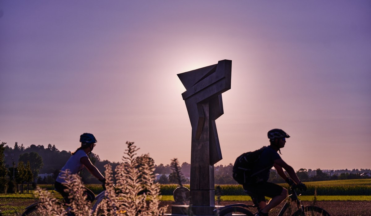 Zwei Radfahrer fahren an einer modernen Skulptur vorbei, die von der untergehenden Sonne beleuchtet wird. Der Himmel ist violett gefärbt., © Natur.Nah. Schönbuch & Heckengäu Zwei Radfahrer fahren an einer modernen Skulptur vorbei, die von der untergehenden Sonne beleuchtet wird. Der Himmel ist violett gefärbt., © Natur.Nah. Schönbuch & Heckengäu