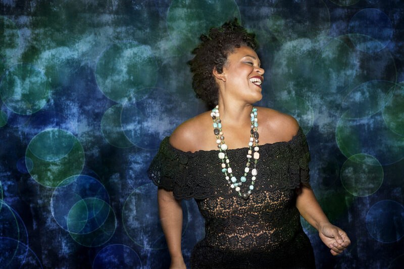 Frau in schwarzem Spitzenkleid und bunter Halskette lächelt vor einem blau-grünen, abstrakten Hintergrund. Sie wirkt fröhlich und entspannt., © Jazzclub Schwäbisch Hall e.V. Frau in schwarzem Spitzenkleid und bunter Halskette lächelt vor einem blau-grünen, abstrakten Hintergrund. Sie wirkt fröhlich und entspannt., © Jazzclub Schwäbisch Hall e.V.