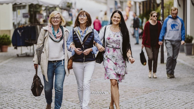 Drei Frauen gehen lachend Arm in Arm durch eine belebte Fußgängerzone in Schwäbisch Hall. Im Hintergrund sind weitere Passanten und Geschäfte zu sehen., © Nico Kurth Drei Frauen gehen lachend Arm in Arm durch eine belebte Fußgängerzone in Schwäbisch Hall. Im Hintergrund sind weitere Passanten und Geschäfte zu sehen., © Nico Kurth